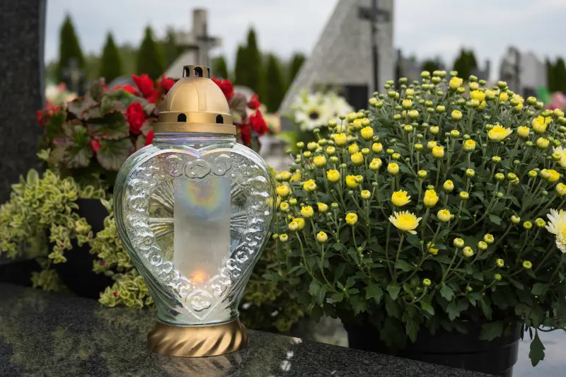 Żółte chryzantemy w doniczce i szklany znicz w kształcie serca stoją na granitowym nagrobku – jesienna dekoracja grobu po Wszystkich Świętych.